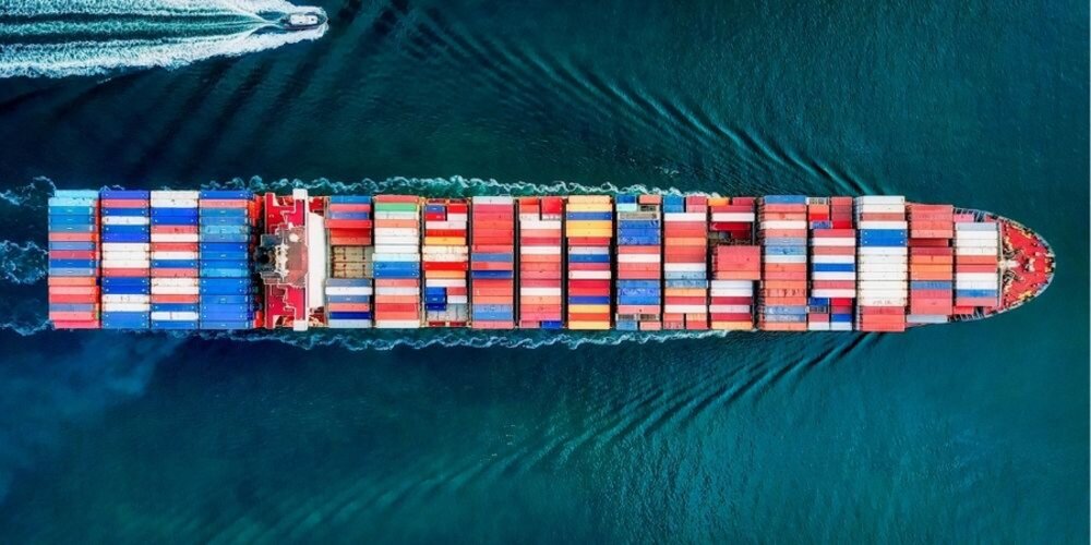 a large fully loaded container ship from above sailing on aqua water flanked by a smaller pilot boat
