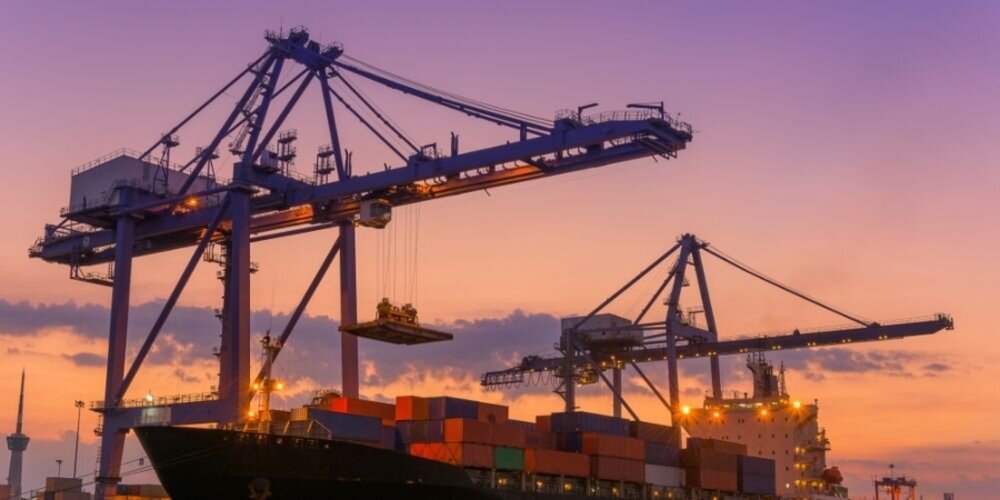 Purple and yellow sunset behind huge port cranes lifting cargo off a large container ship