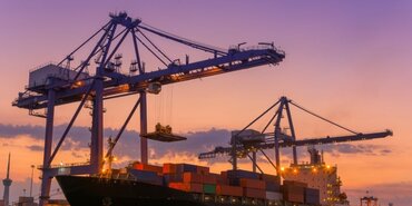 Purple and yellow sunset behind huge port cranes lifting cargo off a large container ship