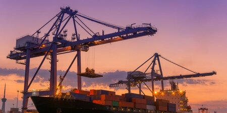 Purple and yellow sunset behind huge port cranes lifting cargo off a large container ship