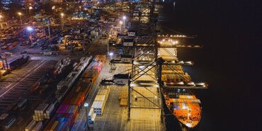 Cargo ship docked alongside port from above. It is nighttime and the sea to the right of the ship appears black. The port on the right hand side is brightly lit.