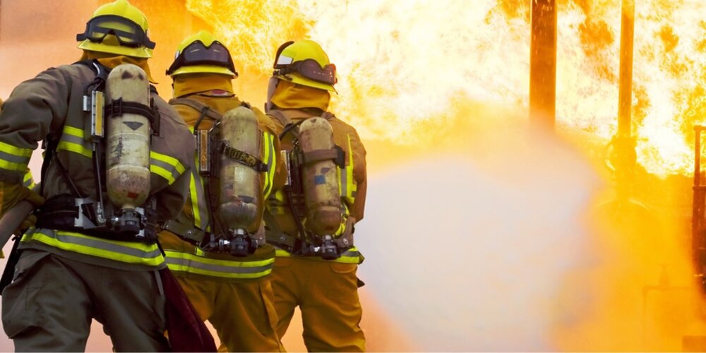 Three firefighters tackle a blaze.The image is taken behind the three firefighters who are spraying the fire with a hose.