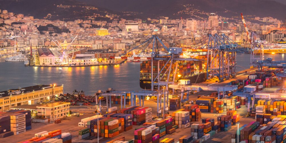 The Port of Barcelona at night. Mountains sit in darkness behind a glittering port. The glow from the lighting lends a yellow-golden hue to the busy scene of loading and unloading cargo