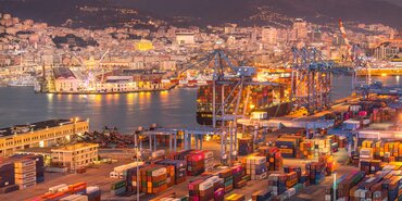 The Port of Barcelona at night. Mountains sit in darkness behind a glittering port. The glow from the lighting lends a yellow-golden hue to the busy scene of loading and unloading cargo