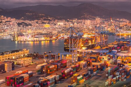 The Port of Barcelona at night. Mountains sit in darkness behind a glittering port. The glow from the lighting lends a yellow-golden hue to the busy scene of loading and unloading cargo