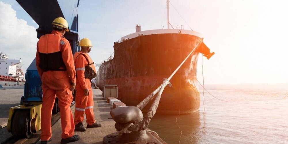 men in hard hats stand near a bollard with a ship tied to it by a large rope and chain